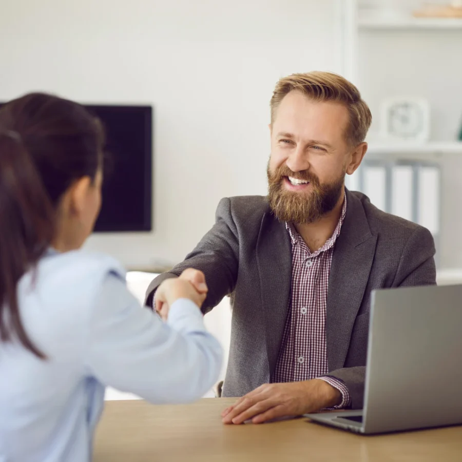 A smiling man with a beard shakes hands with a woman across a desk with a laptop.